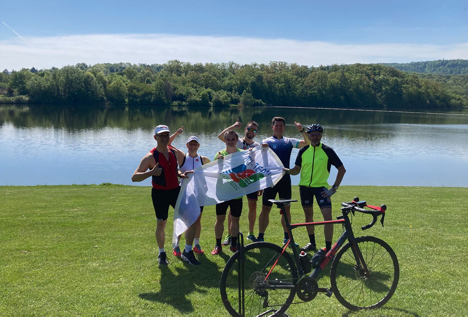 Triathleten am See nach Radtraining mit Vereinsflagge bei Sonnenschein Sportgruppe mit Fahrrad am Flussufer, präsentiert eine weiße Flagge bei sonnigem Wetter