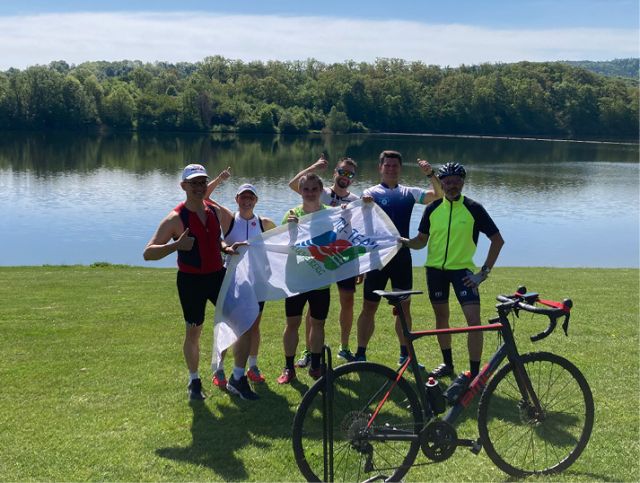 Sportgruppe mit Fahrrad am Flussufer, präsentiert eine weiße Vereinsflagge